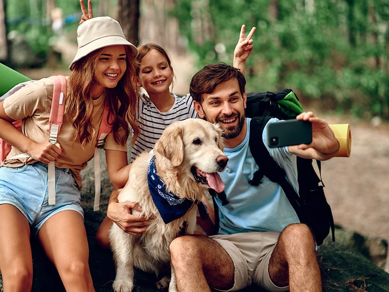 A imagem mostra uma família feliz em um momento de lazer ao ar livre, composta por uma mulher, um homem e uma menina, todos sorrindo enquanto tiram uma selfie. O cachorro da raça Golden Retriever, também parte da família, aparece no centro da foto com uma expressão alegre. O cenário sugere que eles estão desfrutando de uma aventura juntos, destacando o prazer de viajar com pets.